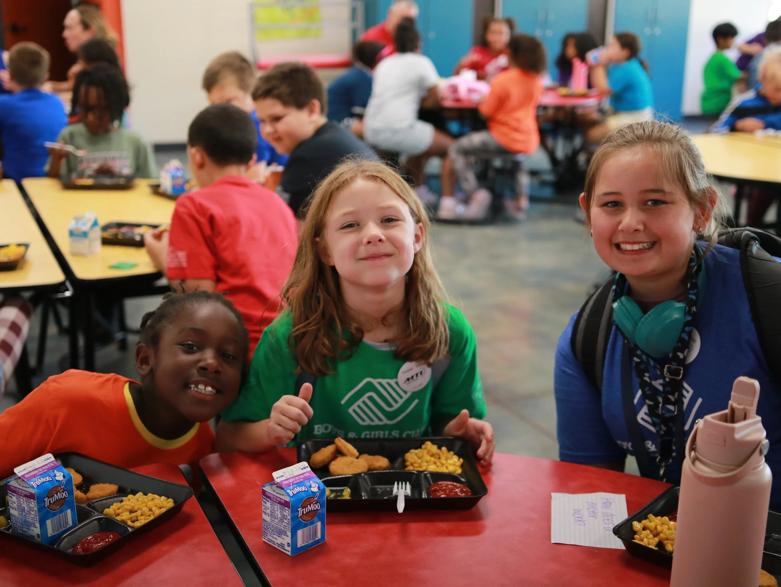 Club members smiling at a lunch table with trays of chicken nuggets, corn and milk
