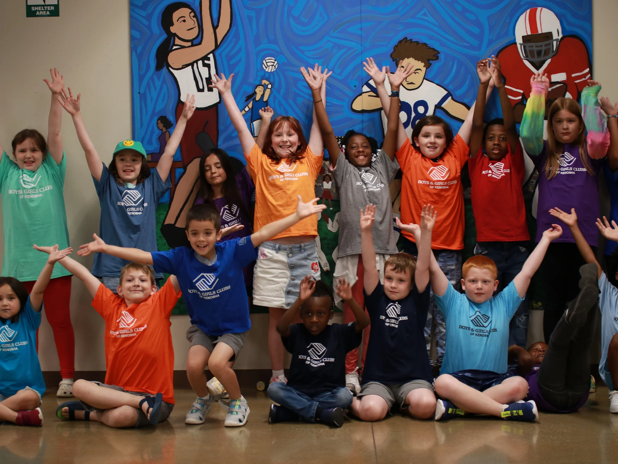 Hero Image A large group of Club members cheering with arms raised in front of a sports mural