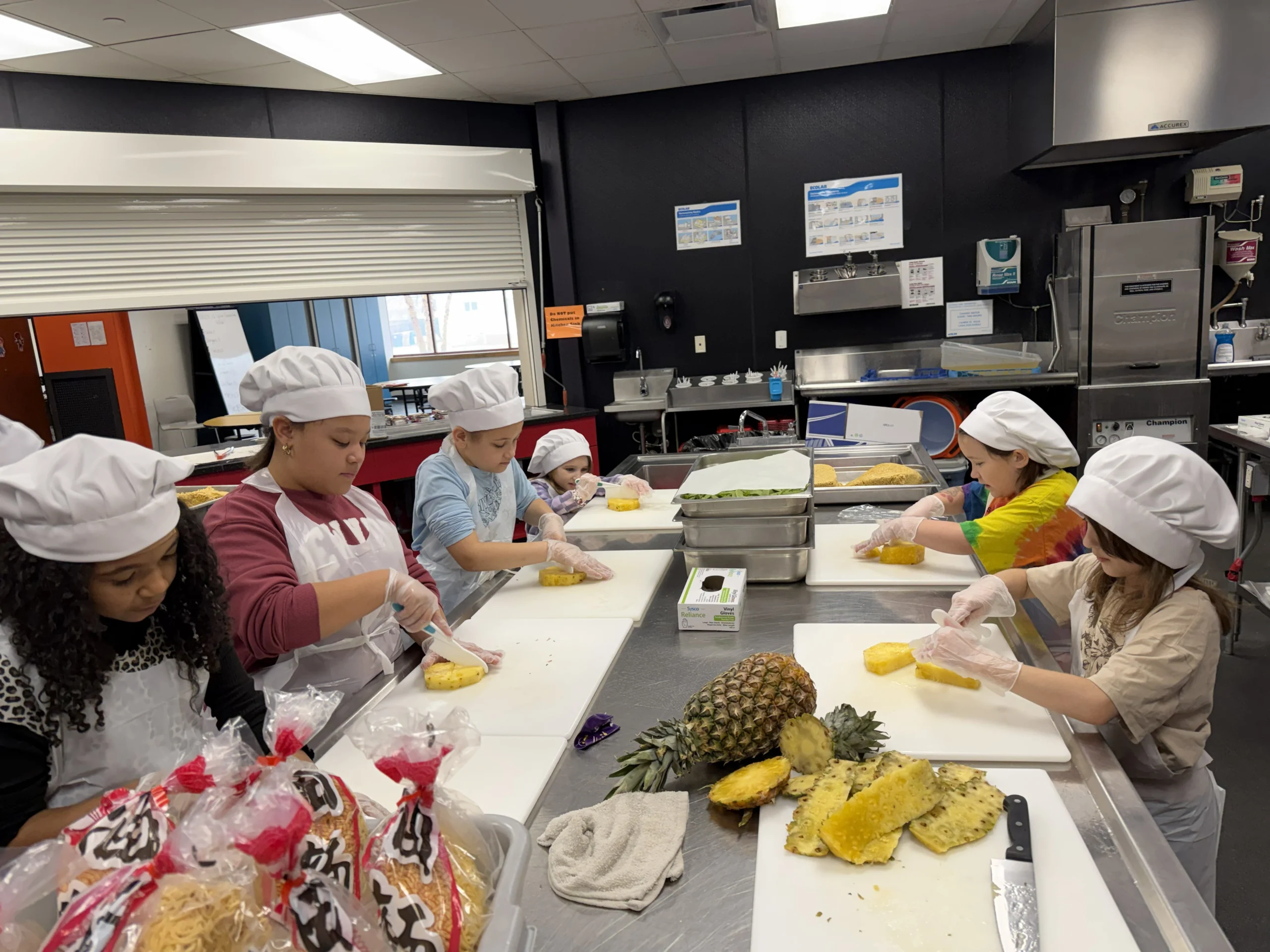 Img 20260306 123239 Club members in chef hats and aprons cutting fresh pineapple in the Club kitchen