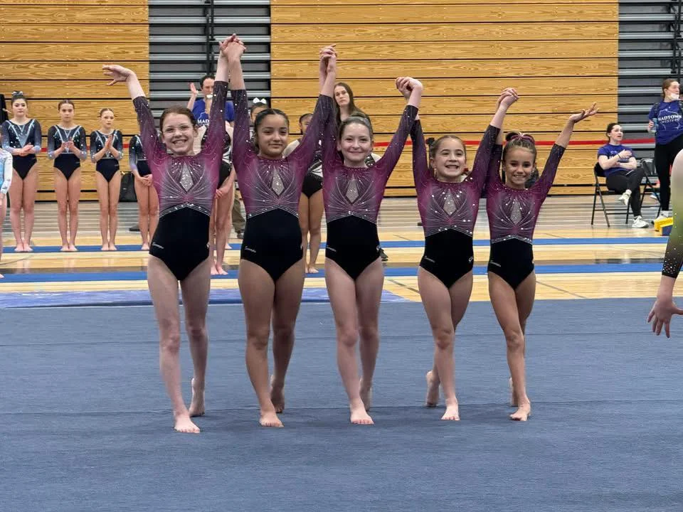 Five gymnasts in matching team leotards raising joined hands on the floor at a meet