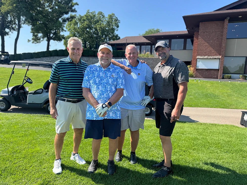 2026 Golf Outing 1 Four golfers in polos and caps posing in front of the clubhouse at the 2026 Golf Outing